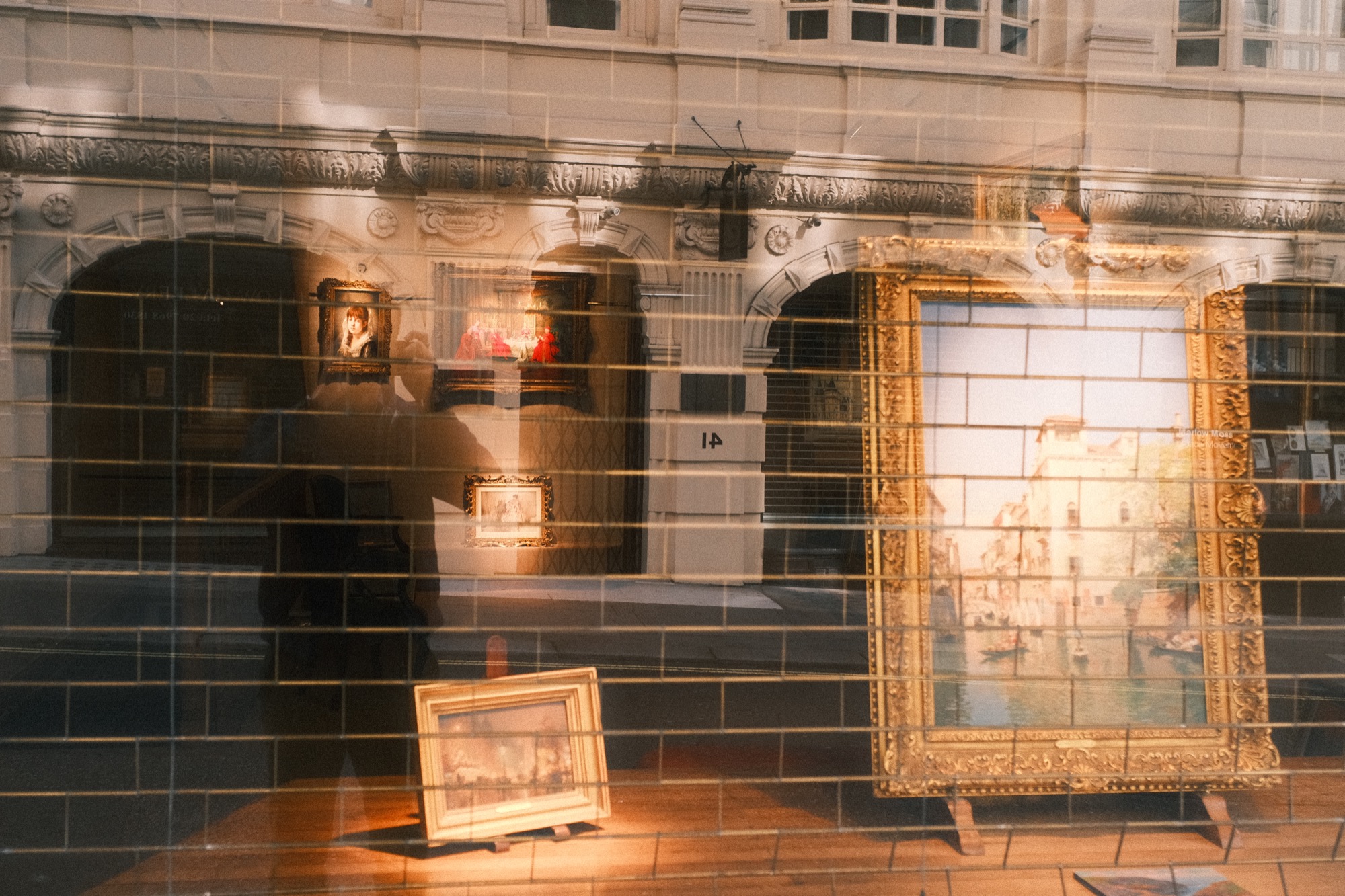 A gallery window where the street is reflected over a gilt-framed painting.