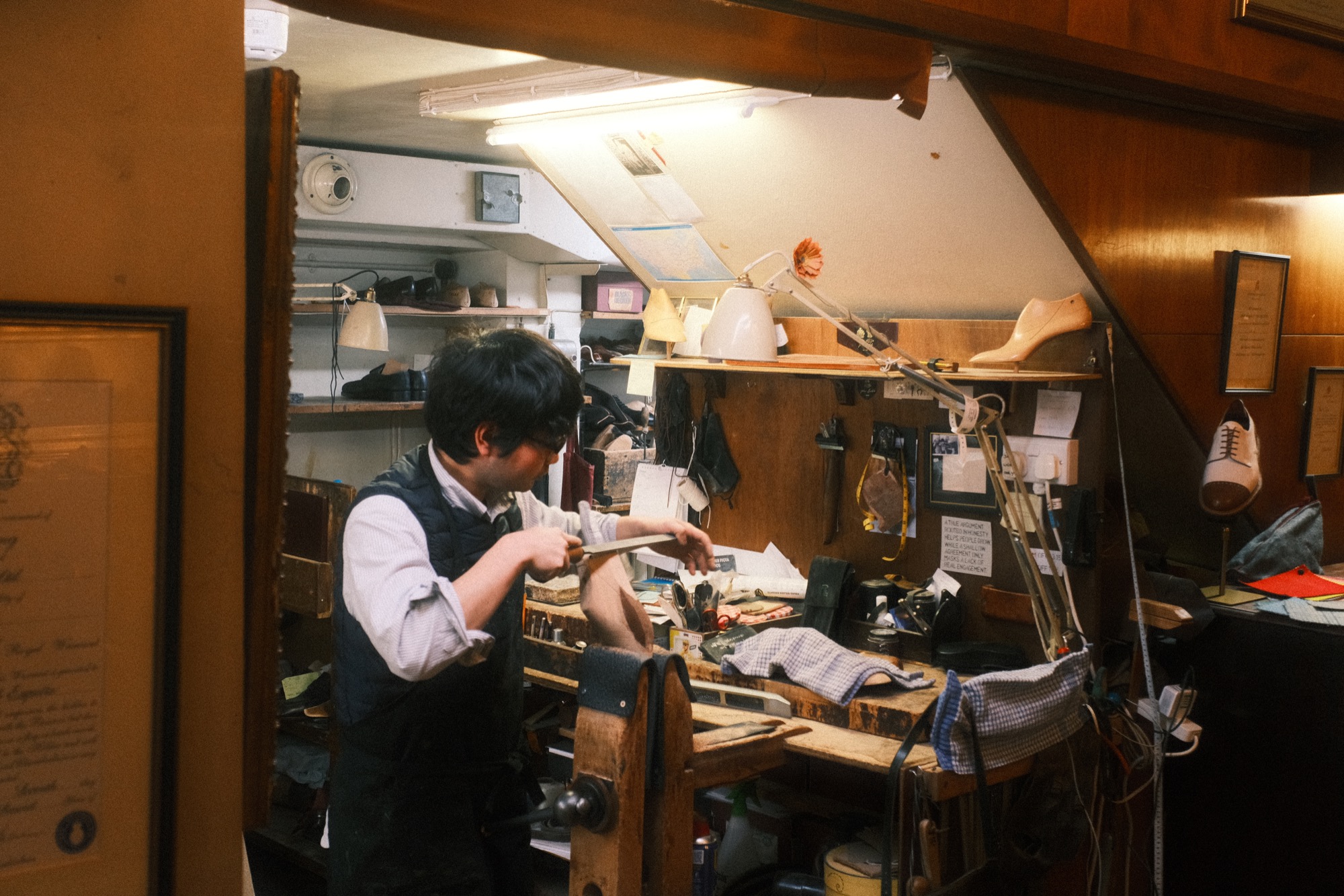 The John Lobb workshop, with a man shaping a wooden last at a bench under a sloped ceiling.
