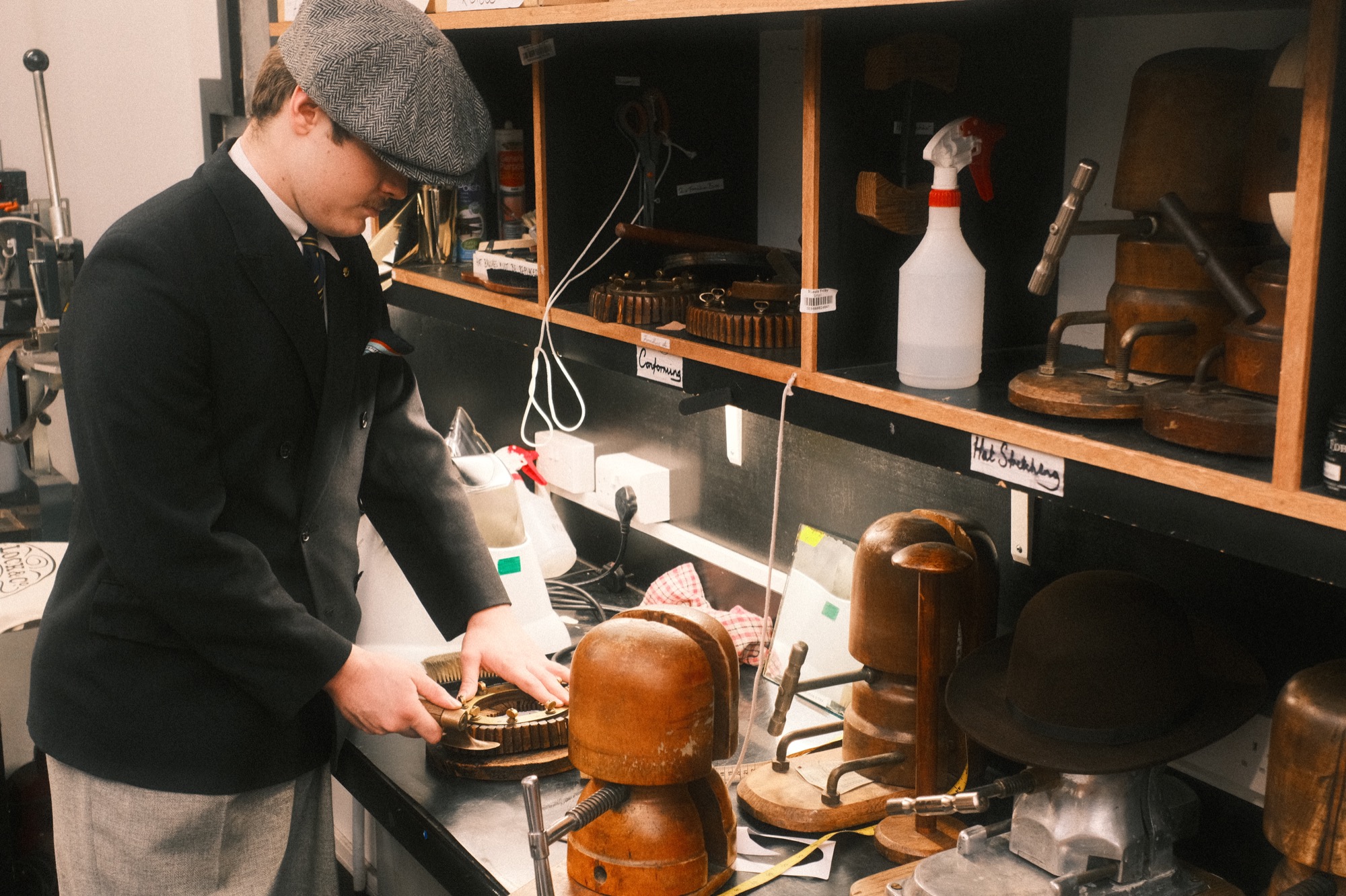 James Anderson in the Lock & Co. hat shop, in a light grey hat, with wooden hat-making tools on the shelves.